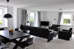 Ein modernes Wohnzimmer mit einer schwarzen Couch, einem Esstisch, Stühlen, großen Fenstern mit Vorhängen und einem Flachbildfernseher.