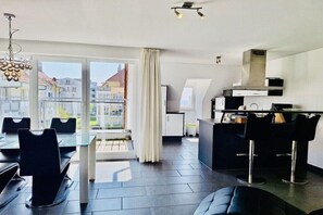 Et moderne køkken med komfur, mikroovn og skabe, et spiseområde med sorte stole og et glasbord, samt store vinduer med udsigt til bygninger.