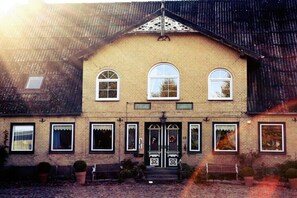 A two-story building with a symmetrical facade, a central entrance with a door and windows, and a small porch area with benches.