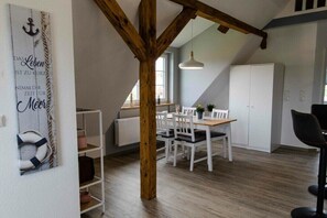 A dining area with a wooden beam ceiling, white chairs, a table, and a white cabinet. - Willi's view of the meadow (Grödersby)