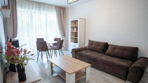 Una sala de estar con un sofá, una mesa de centro, una estantería, una zona de comedor con sillas y una ventana con cortinas.