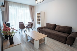 A living room with a sofa, a coffee table, a bookshelf, a dining area with chairs, and a window with curtains.
