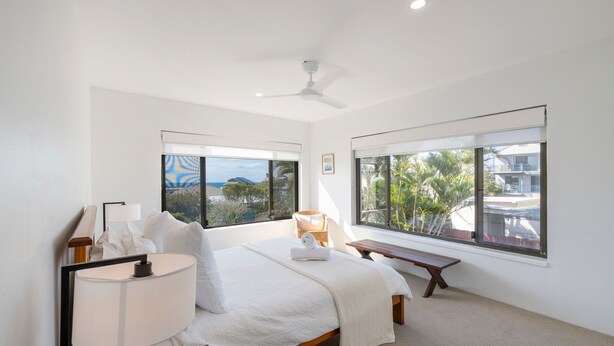 Un dormitorio con cama, una banca, un escritorio y dos ventanas con vistas a palmeras y edificios.