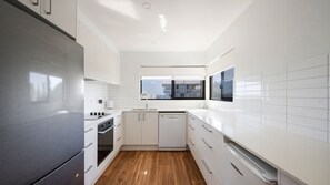 A modern kitchen with white cabinetry, a stainless steel refrigerator, a dishwasher, and a window providing a view of the outside. - Talinga 4 - Lifestyle Yamba (YAMBA)