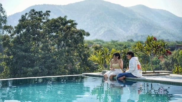 Two people sitting by a pool with a mountainous backdrop. - The Riverside Estate (Udaipur)