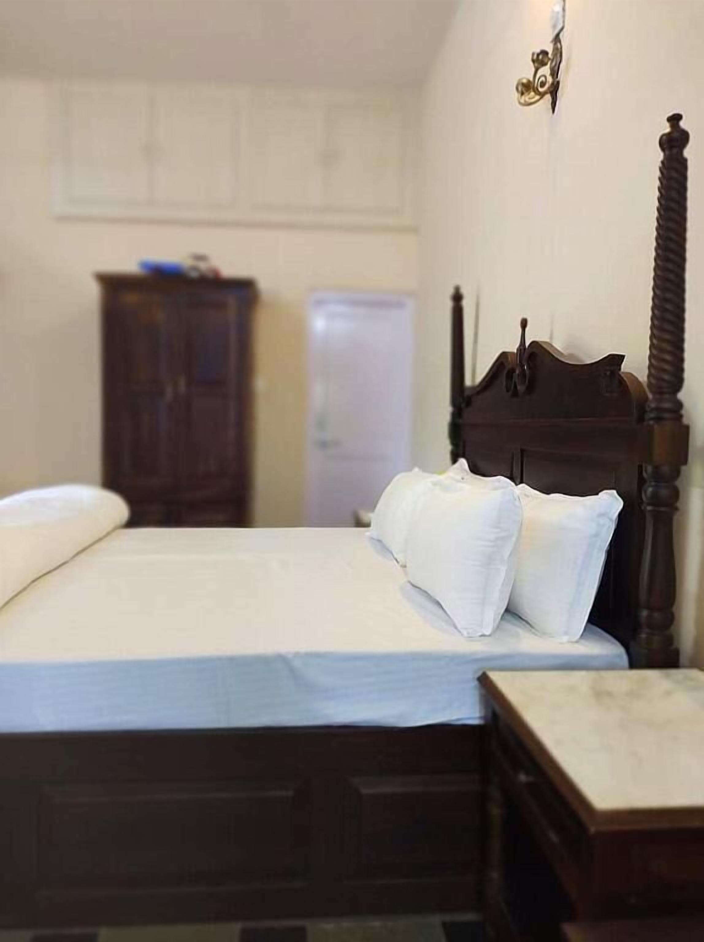 Een slaapkamer met een houten hemelbed, een nachtkastje en een kledingkast op de achtergrond.