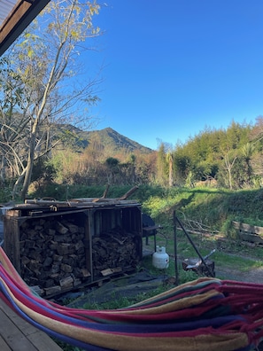 Una cadira d'hamaca, una estructura de fusta amb una pila de llenya i un cilindre de gas en un entorn natural a l'aire lliure, amb una muntanya al fons.