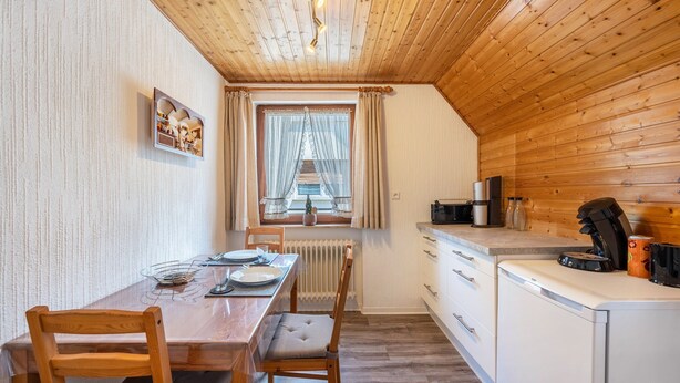 A wooden interior with a dining table, chairs, a kitchen area, and a window with curtains.
