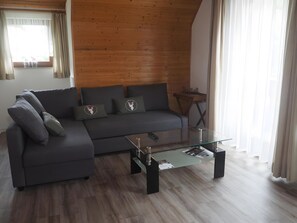 A modern living room with a dark grey sectional sofa, a glass coffee table, wooden flooring, and a wooden ceiling.