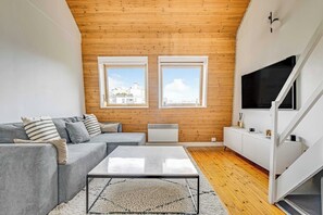 Ein modernes Wohnzimmer mit grauem Sofa, einem Couchtisch und einem weißen Schrank. Zwei Fenster bieten Blick auf Gebäude, die Decke ist aus Holz.