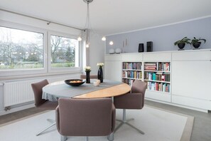 A modern dining area with a table, chairs, a bookshelf, and a view through large windows. - Holiday home with 3 bedrooms (Ostseebad Binz)
