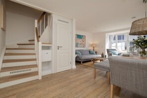 A staircase with wooden steps and white walls, a gray sofa with blue cushions, a dining table with chairs, and a hanging light fixture. - Holiday home in Hörnum with swimming pool (Hörnum)