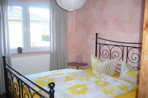 Ein Schlafzimmer mit Bett, Fenster, Heizkörper, kleinem Tisch und einer Topfpflanze.