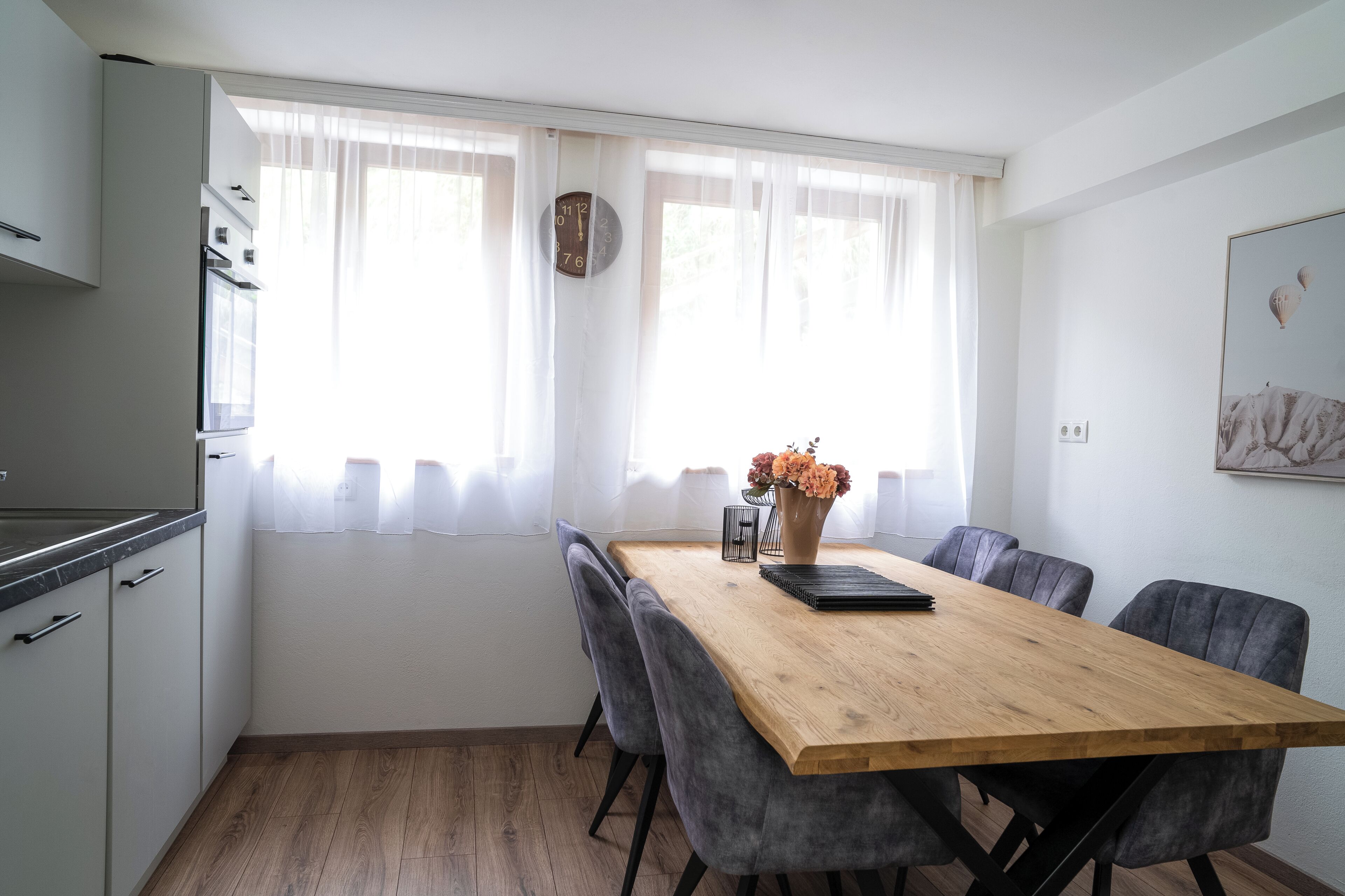 Una cocina moderna con armarios blancos, una mesa de comedor de madera con sillas grises y un reloj en la pared.