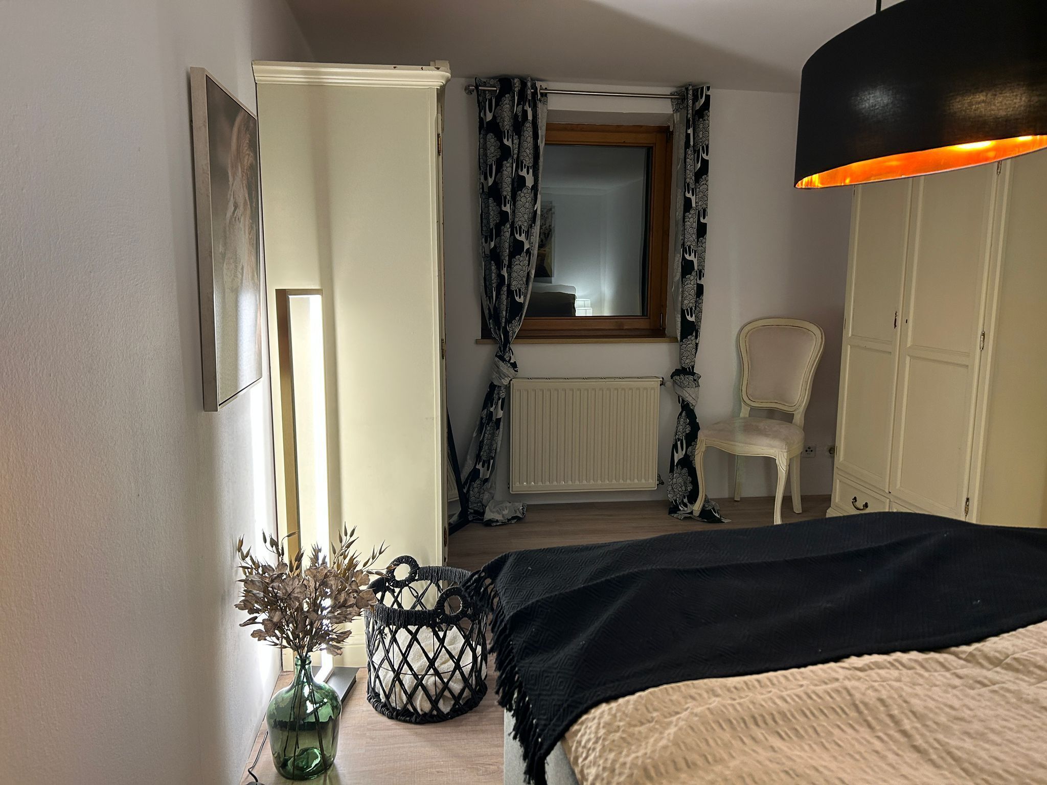 A bedroom with a bed, a chair, a radiator, a window with curtains, and a hanging light fixture.