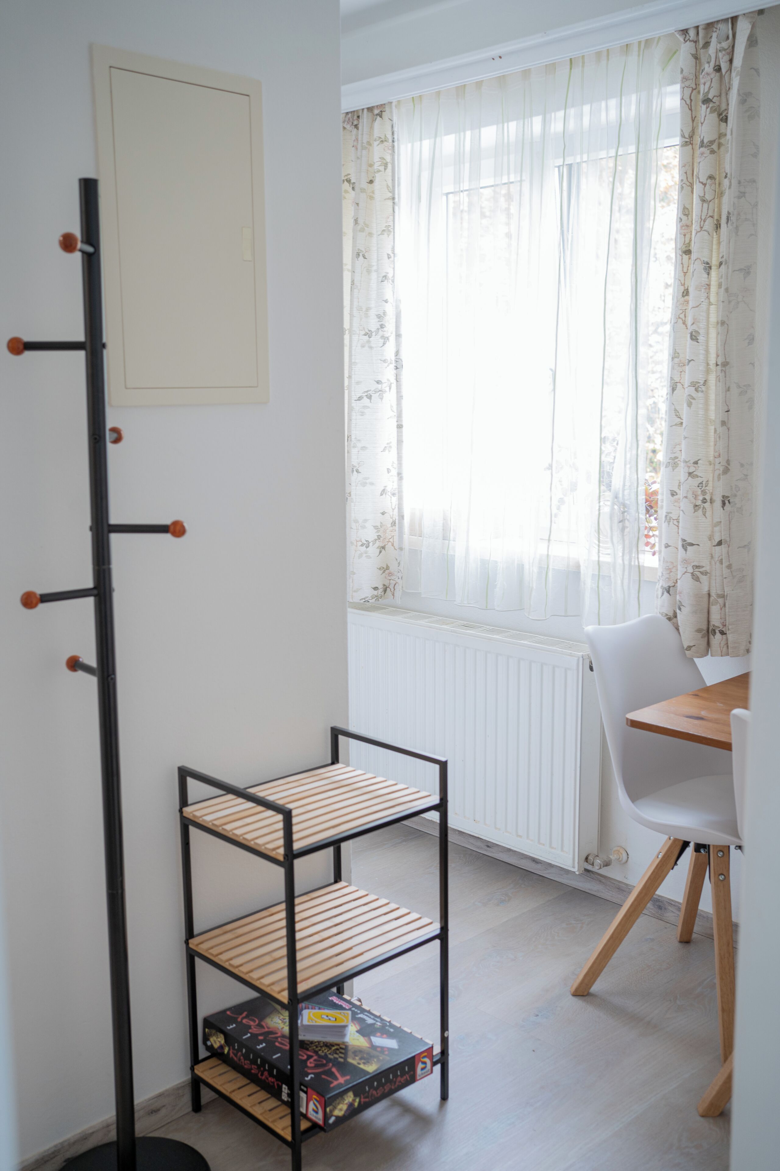 Una habitación con perchero, radiador blanco, ventana con cortinas florales y mesa de comedor con una silla.