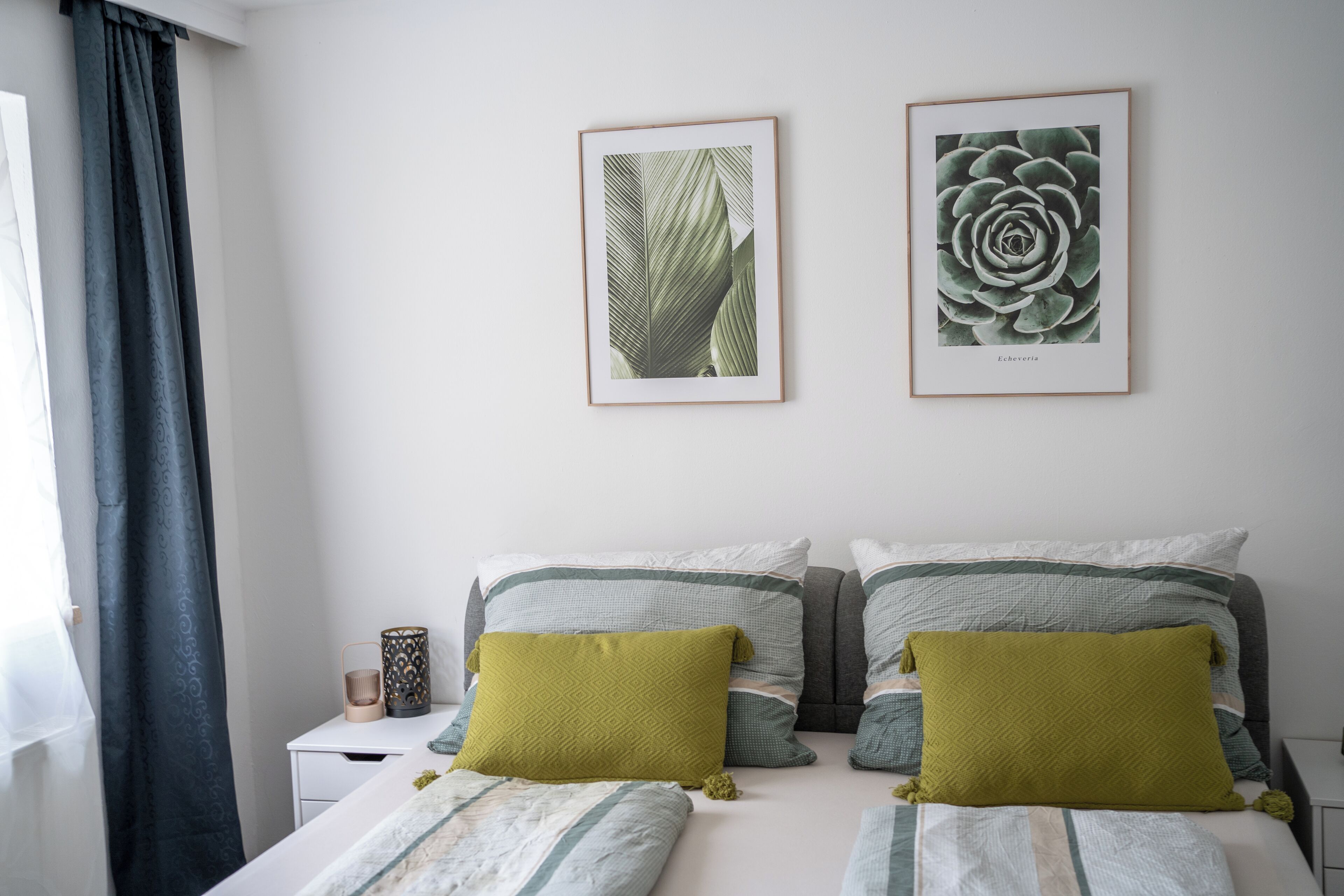A bedroom with a bed, two framed botanical prints on the wall, and a nightstand with a lamp.
