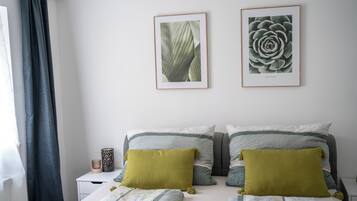 A bedroom with a bed, two framed botanical prints on the wall, and a nightstand with a lamp.