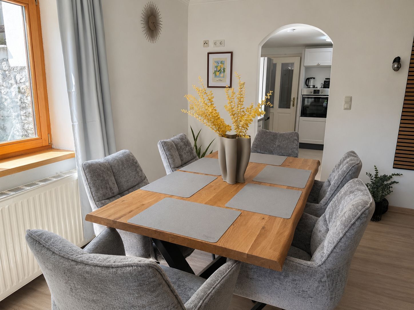 Un comedor con una mesa de madera, sillas tapizadas en gris y un jarrón con flores amarillas.