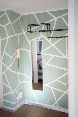 Ein Zimmer mit Wand im geometrischen Muster, einem spiegelnden, weiß umrandeten Spiegel, einem Telefon und einem schwarzen Metallregal.