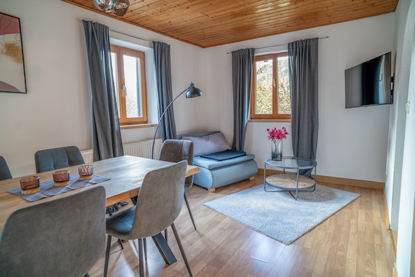 Ein modernes Zimmer mit Holzboden, einem Esstisch mit Stühlen, einer Couch, einem an der Wand befestigten Fernseher und einem Fenster mit Vorhängen.