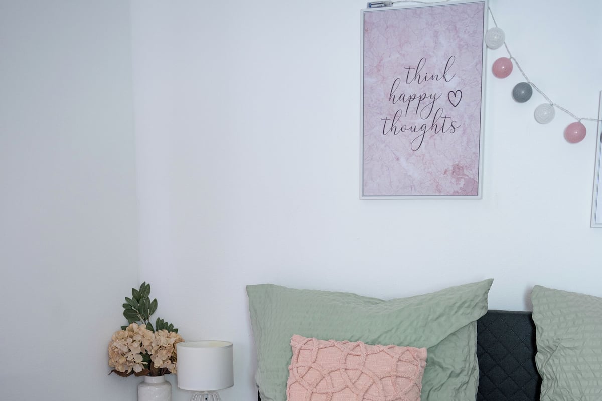A neatly arranged bedroom with a bed, pillows, a bedside table with a lamp, and a wall-mounted sign.