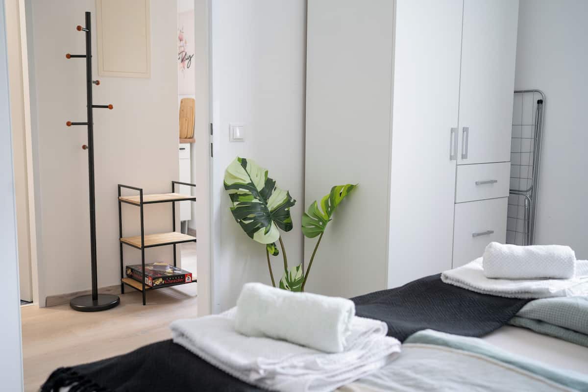 A hotel room with a bed, a coat rack, a shelf with books, and a large plant.