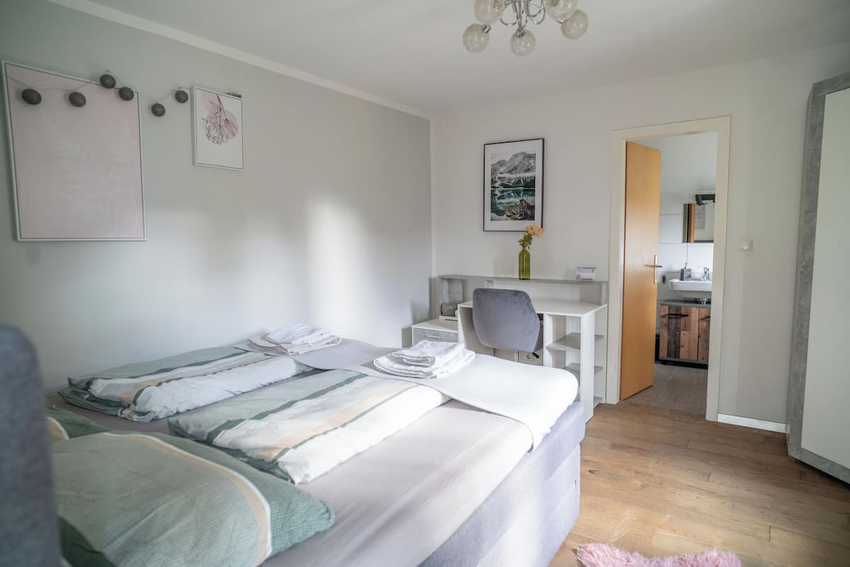 A bedroom with a bed, a desk, and a chair. There is a bathroom visible through an open door.