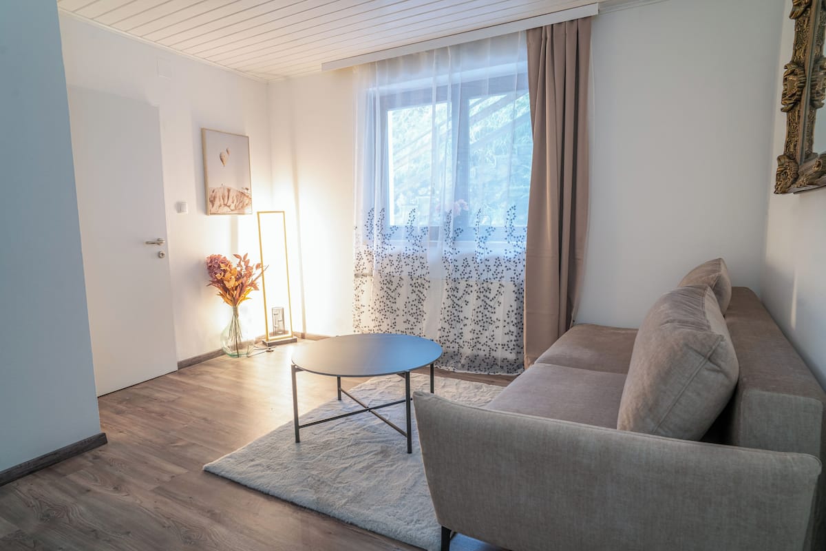 A living room with a sofa, a small round table, a vase with flowers, and a window with curtains.
