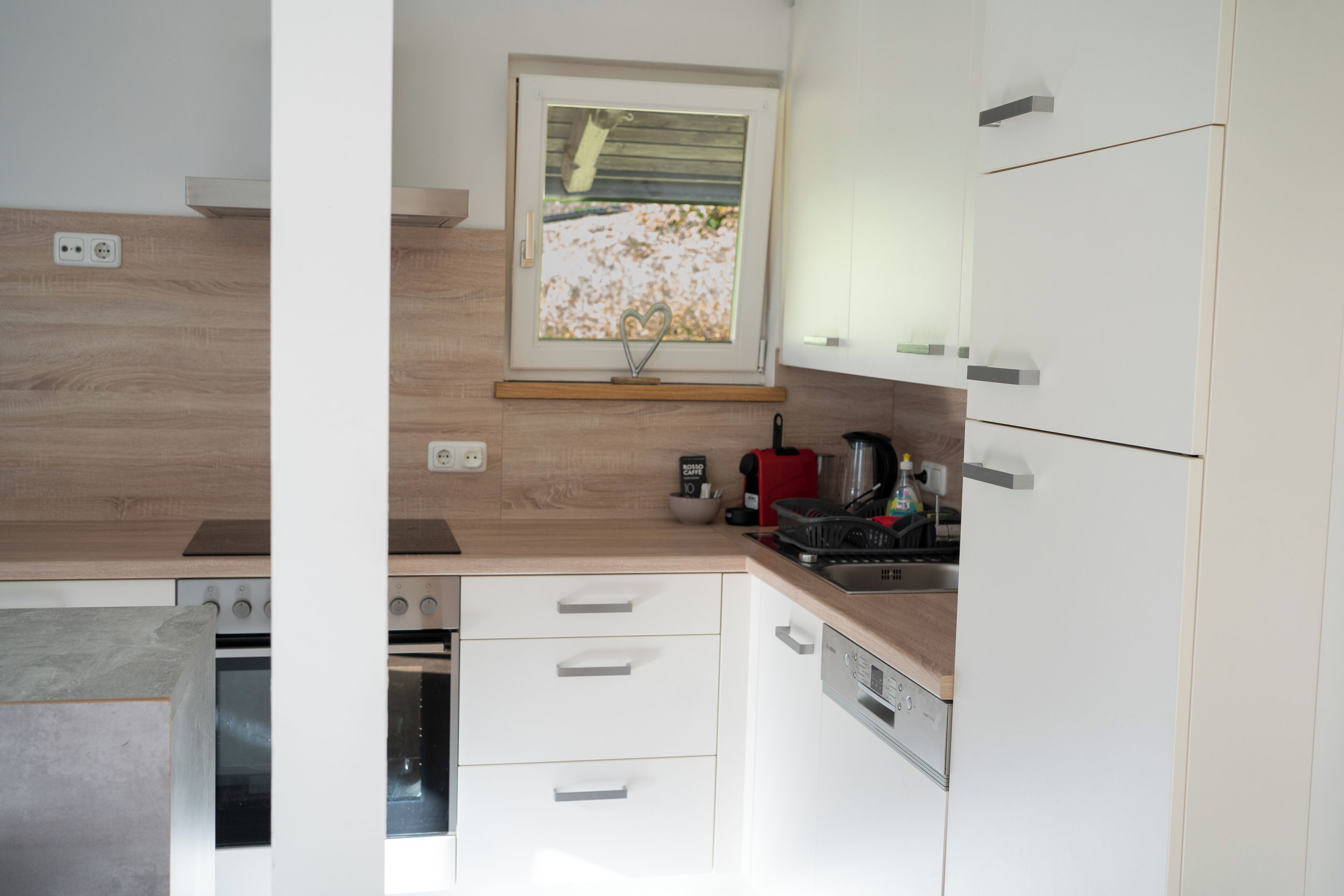 A modern kitchen with white cabinets, a wooden countertop, a built-in oven, a dishwasher, and a window with a view of a garden.