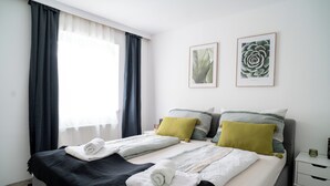 A neatly made bed with rolled towels, a bedside table with a vase, and two framed botanical prints on the wall.