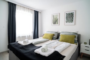 Ein ordentlich bezogenes Bett mit aufgerollten Handtüchern, ein Nachttisch mit einer Vase und zwei gerahmte Pflanzenbilder an der Wand.