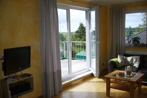 Ein Wohnzimmer mit Fernseher, einem Glastisch, einer Couch und einer Schiebetür, die auf einen Balkon führt.
