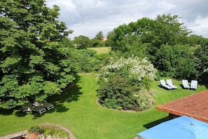 Ein üppiger Garten mit grünen Rasenflächen, Bäumen und einer Terrasse mit Liegestühlen und einem Tisch.
