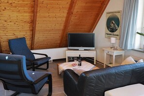 Ein gemütliches Zimmer mit schrägen Wänden, einem Fernseher auf einem Standfuß, einem Sofa, einem Sessel und einem kleinen Tisch mit einer Lampe.