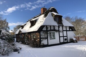 Ein traditionelles Fachwerkhäuschen mit einem steilen Walmdach, umgeben von schneebedeckten Bäumen und blauem Himmel.