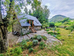 A stone house with a sloped roof, wooden steps leading to a door, and a garden with various plants and a barbecue area. - Cozy studio on the first floor of a mountain barn (Ercé)