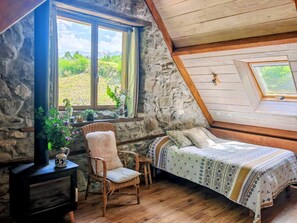 Ein gemütliches Zimmer mit einem Bett, einem Sessel, einem kleinen Tisch mit einer Vase und einem Fenster mit Blick auf Grünflächen.
