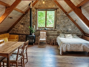 Ein gemütliches Schlafzimmer im Dachgeschoss mit Steinwand, Holzbalkendecke, einem Bett mit gemusterten Textilien, einem kleinen Tisch und einem Stuhl.