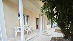 A covered outdoor seating area with a table and chairs, a door, and a small garden area with a tree. - Quiet, Modern Ground-floor Apartment in Lignano (Lignano Sabbiadoro)