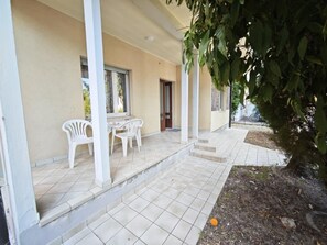 Ein überdachter Außenbereich mit Tisch und Stühlen, einer Tür und einem kleinen Garten mit einem Baum.