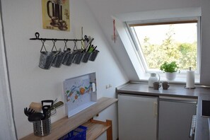 Et køkken med et vindue, en metalkurv med redskaber, et indrammet billede og en lille plante.