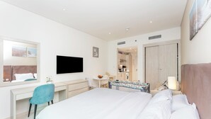 A modern hotel room with a large bed, a desk with a chair, a television, a small table with a lamp, and a view of the kitchen. - Modern studio in Seven Palm, pool and beach (Dubai)