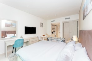 Ein modernes Hotelzimmer mit einem großen Bett, einem Schreibtisch mit Stuhl, einem Fernseher, einem kleinen Tisch mit Lampe und Blick auf die Küche.