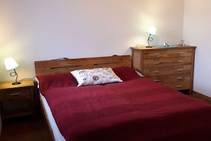 Quarto com cama de madeira, manta vermelha, mesa de cabeceira com abajur e cômoda com espelho.