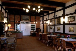 Uma hospedaria tradicional com vigas de madeira aparentes, lareira de tijolos e área de refeições com mesas e cadeiras.