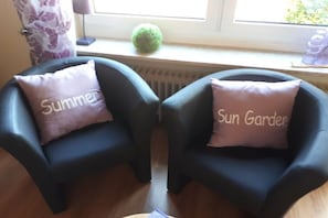 Zwei schwarze Sessel mit Kissen mit den Aufschriften 'Summer' und 'Sun Garden', eine Heizung, ein grüner, kugelförmiger Gegenstand und ein Fenster mit Vorhängen.