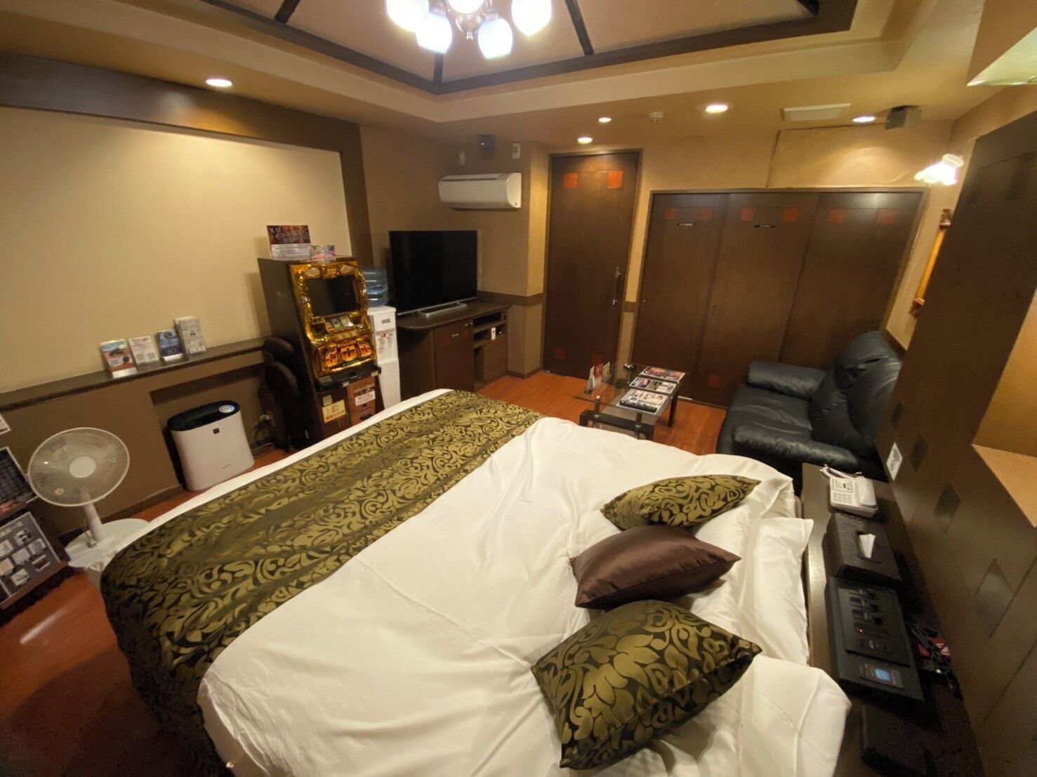 A hotel room with a bed, a television, a desk with a computer, a fan, and a door.