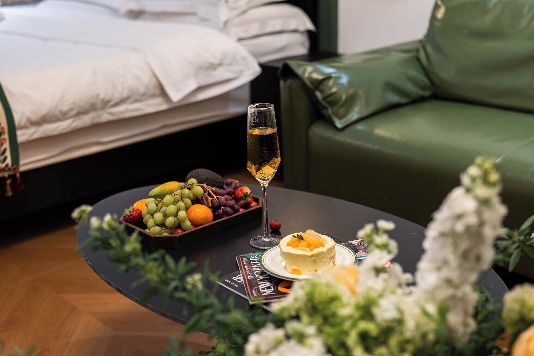 Quarto bem arrumado com cama, um sofá verde, mesa com uma cesta de frutas, uma taça de champanhe e uma revista.