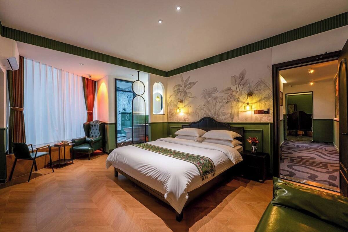 A hotel room with a large bed, a sitting area with a chair and table, a mirror, and a view of the outdoors.